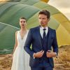Detailaufnahme eines Bräutigams im blauen Hochzeitsanzug mit taillierter Silhouette und strukturierter Weste. Im Hintergrund ist ein Brautkleid.