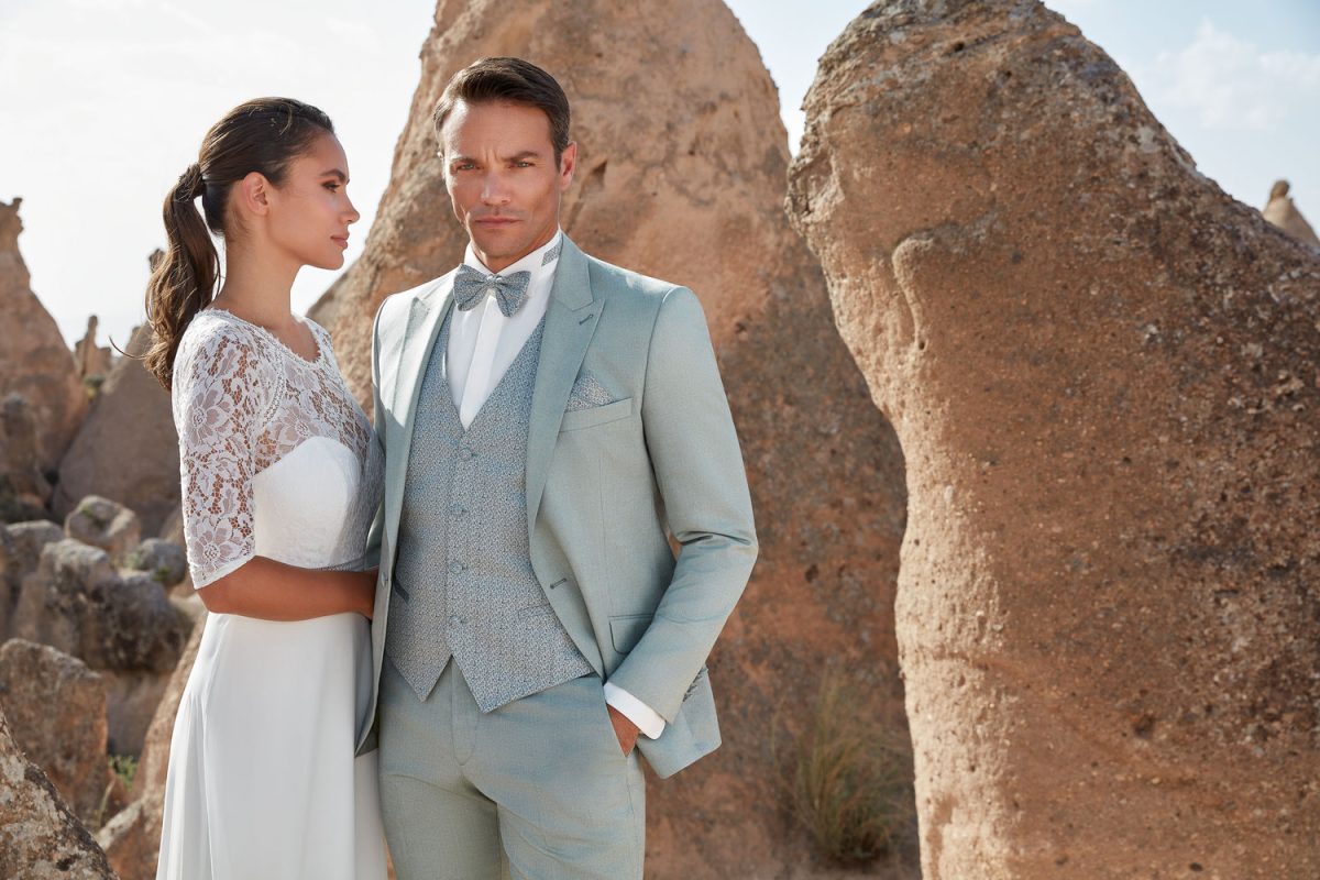 Detailaufnahme eines hellgrünen Hochzeitsanzugs mit schmalem Revers, Weste und weißem Hemd und eines Brautkleides mit Spitzenoberteil