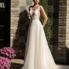 Braut in einem eleganten A-Linie Brautkleid mit tiefem V-Ausschnitt, floraler Tattoo-Spitze und transparentem Tüllrock, stehend vor einer Steinwand im Tageslicht, romantischer Look mit natürlichem Lichtspiel.