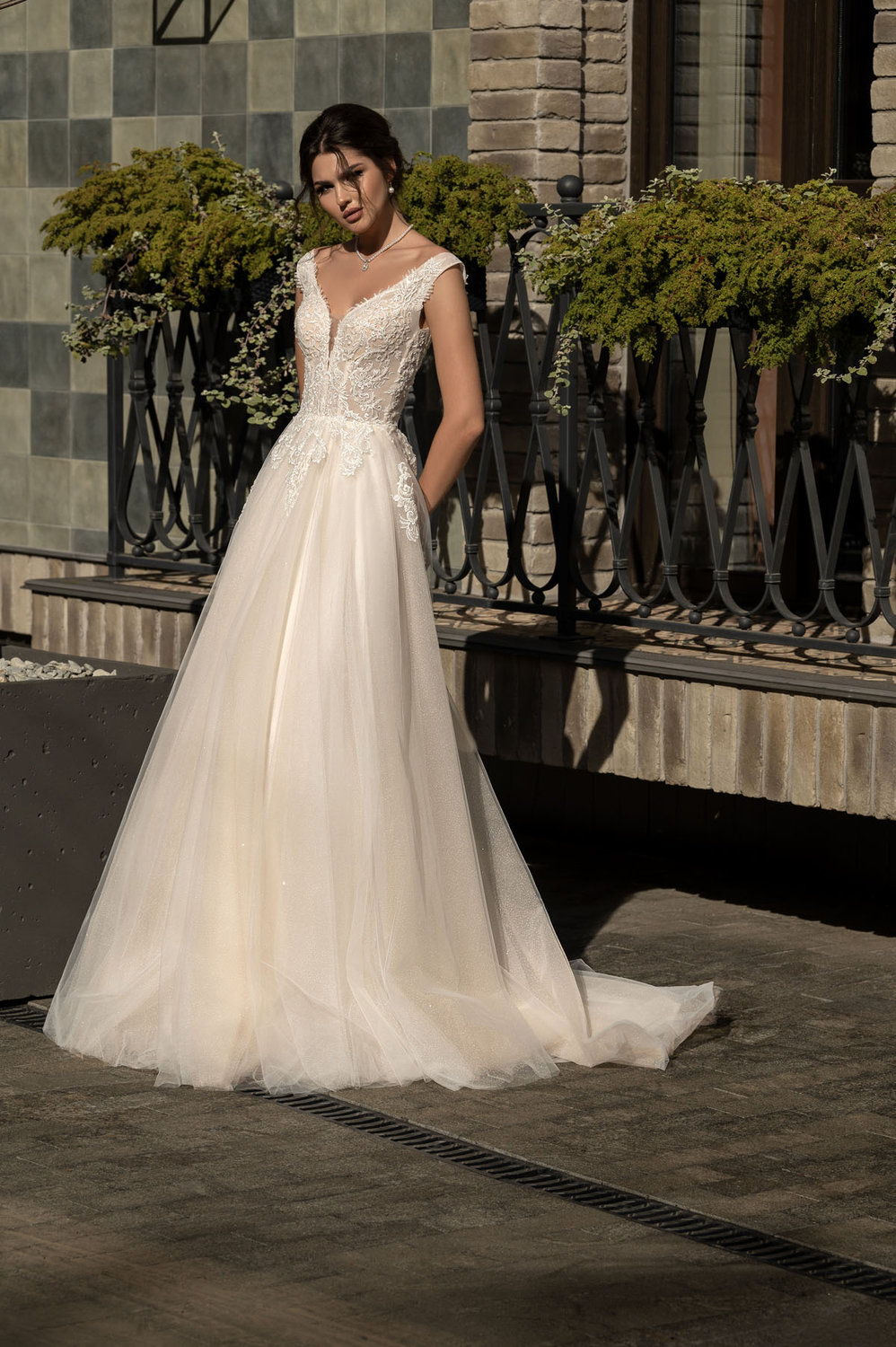 Vollansicht eines eleganten A-Linie Brautkleids mit tiefem V-Ausschnitt, zartem Spitzentop, fließendem Tüllrock und tiefem Rückenausschnitt – fotografiert im Tageslicht vor stilvoller Kulisse.