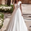 Braut in einem eleganten Brautkleid mit floraler Spitze, V-Ausschnitt und fließendem Tüllrock, stehend in sonnigem Außenbereich vor Blumenbeeten