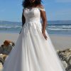 Braut in einem A-Linien-Brautkleid mit Carmen-Ausschnitt und floraler Spitze, stehend am Strand vor dem Meer unter blauem Himmel.
