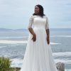 Braut in einem eleganten, langärmligen A-Linien-Brautkleid mit Spitze und fließendem Rock, stehend auf einem Felsen mit Blick auf das Meer.