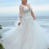 Lachende Braut in einem glitzernden A-Linien-Brautkleid mit tiefem Ausschnitt und Tüllrock, stehend am Meer vor türkisblauem Wasser.