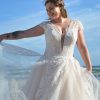 Lächelnde Braut in einem figurbetonten Brautkleid mit tiefem V-Ausschnitt, floraler Spitze und Nude-Schimmer, posierend am Meer unter blauem Himmel.