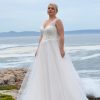 Braut in einem eleganten A-Linien-Brautkleid mit Spitzenträgern und Tüllrock, stehend auf Felsen vor dem Meer bei klarem Himmel.