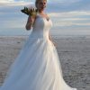 Braut in einem figurbetonten Curvy-Brautkleid mit Spitzenkorsage und fließendem Tüllrock am Strand bei Sonnenuntergang.