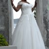Braut in einem romantischen Curvy-Brautkleid mit Spitzenkorsage, Carmen-Ausschnitt und fließendem Tüllrock, stehend zwischen zwei Steinbögen.