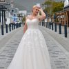 Braut in einem eleganten A-Linien-Brautkleid mit transparentem Illusionsausschnitt und floraler Spitze, posierend auf einer gepflasterten Straße mit Bergkulisse.