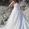 Strahlende Braut in figurbetontem Spitzen-Brautkleid mit V-Ausschnitt am Strand bei Sonnenlicht.