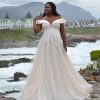 Curvy Braut in einem schulterfreien Brautkleid mit glitzernder A-Linie vor idyllischer Landschaft mit Bergen und Wasser.